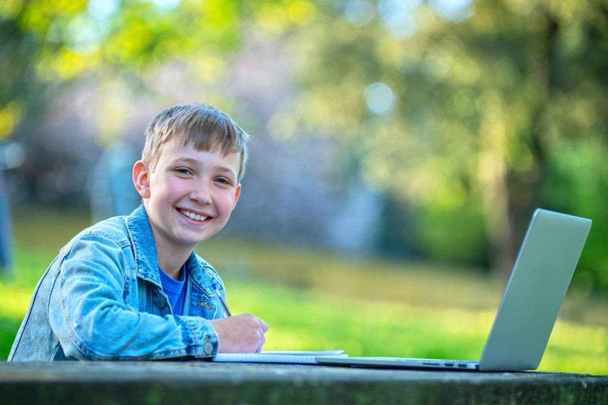 A smiling Year 3 boy studying English outdoors with his laptop and notebook