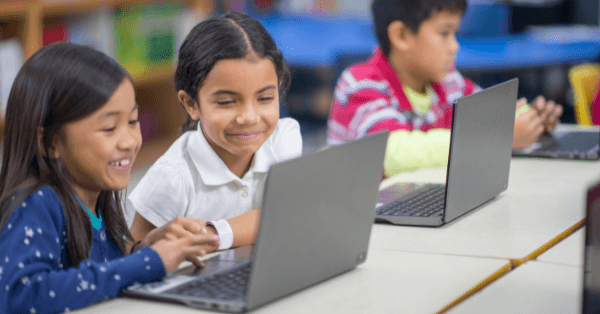 Two smiling girls working together on laptops in a classroom