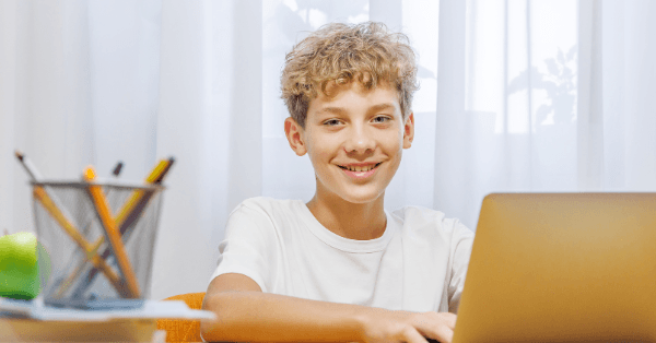 A smiling curly-haired boy sitting at his laptop ready for an online lesson