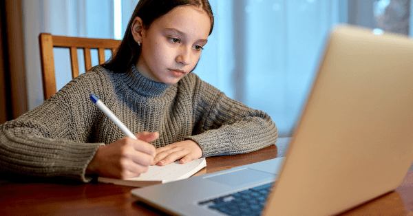 A focused girl writing notes at the table beside her laptop